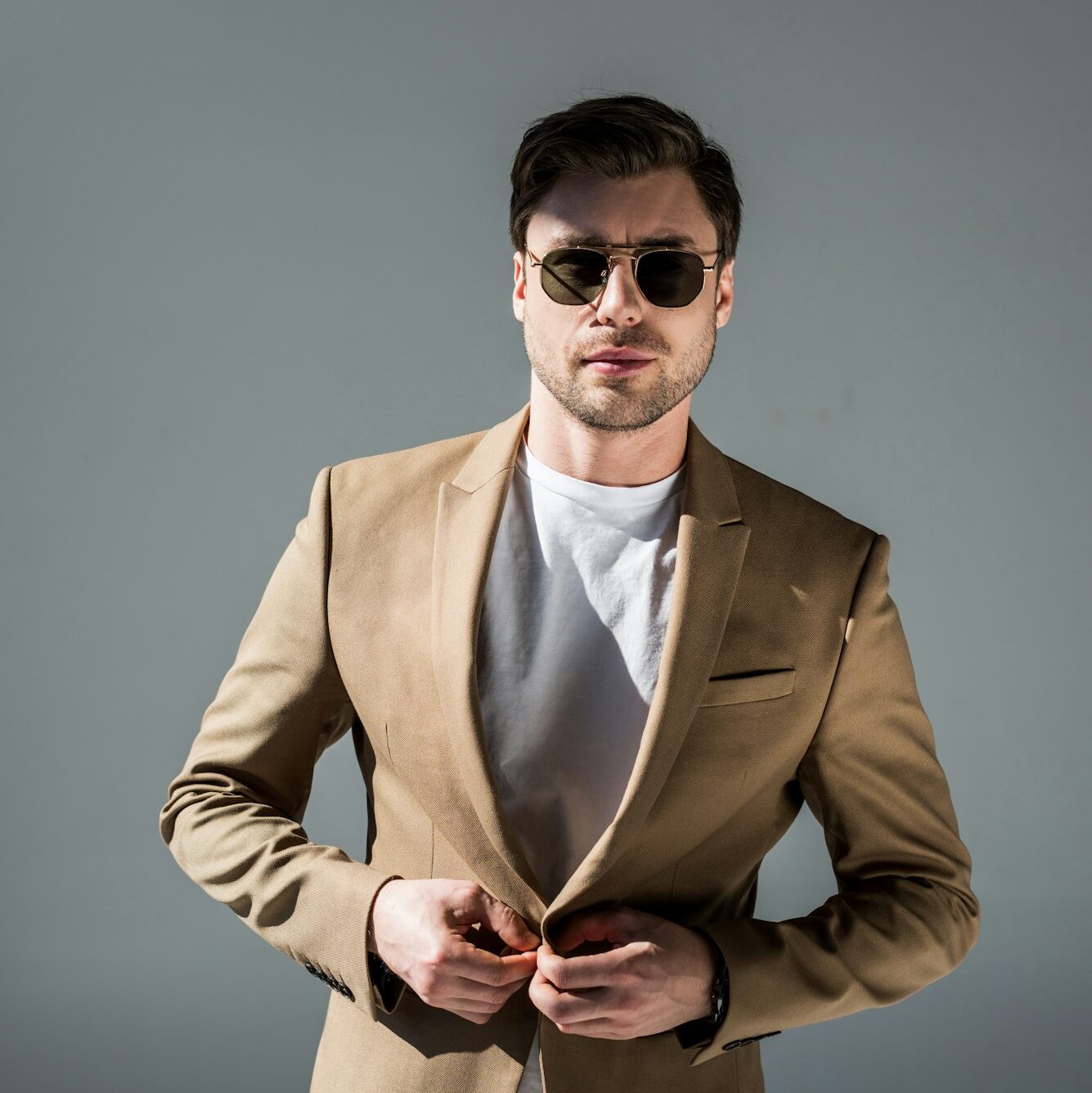 handsome trendy man in beige suite and sunglasses looking at camera on grey